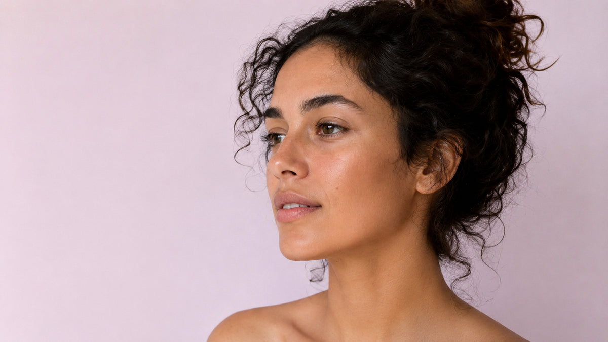 A close-up portrait of a woman with curly dark hair, shown from the shoulders up against a soft light pink background.