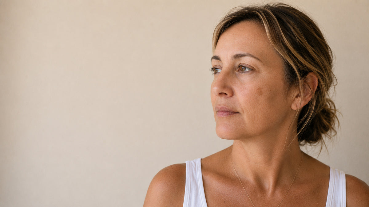A woman with light brown hair tied back, wearing a white tank top, shown in profile against a plain beige wall.