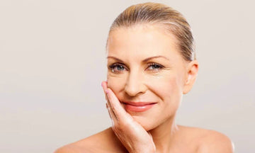 A close-up portrait of a smiling woman with her hand resting on her cheek, set against a plain light gray background.