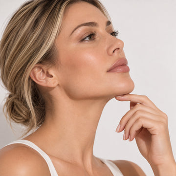 A close-up portrait of a woman with blonde hair in a bun, wearing a white tank top, touching her chin against a clean light gray background.