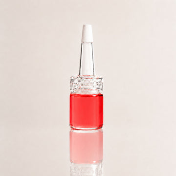 A small clear glass dropper bottle filled with bright red liquid, featuring a clear glass pipette-style top with a white cap, centered on a light beige background with a soft reflection below.