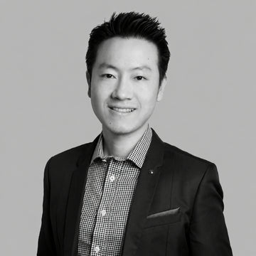 A black and white professional headshot of a smiling man with short dark hair, wearing a dark suit jacket over a small checkered button-down shirt, set against a solid light gray background.