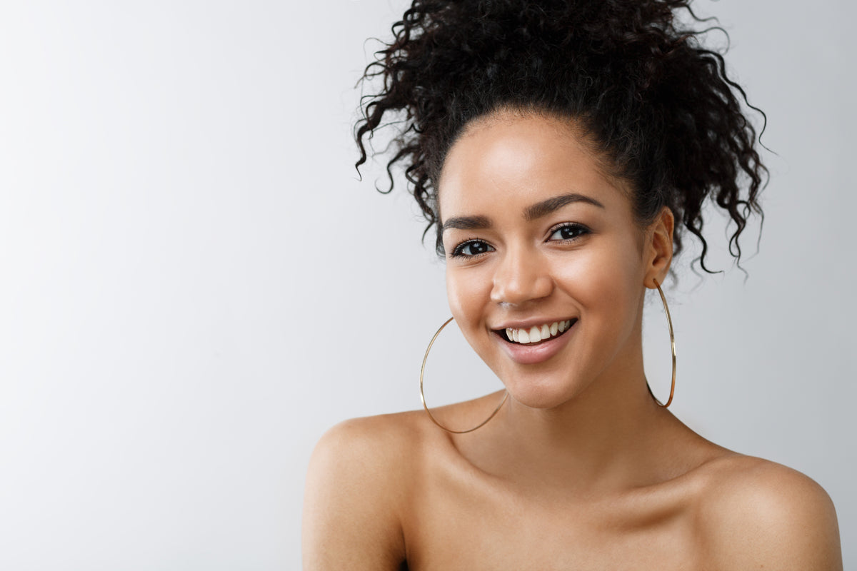 A smiling woman with curly dark hair wearing large gold hoop earrings, photographed against a light gray studio background.