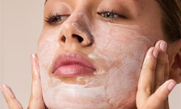 Close-up of a woman's face with a thick white skincare cream or mask applied, with her hands gently touching her cheeks against a plain beige background.