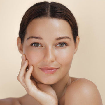 A close-up portrait of a woman with smooth, radiant skin and dark brown hair pulled back, resting her hand on her cheek against a soft beige background.